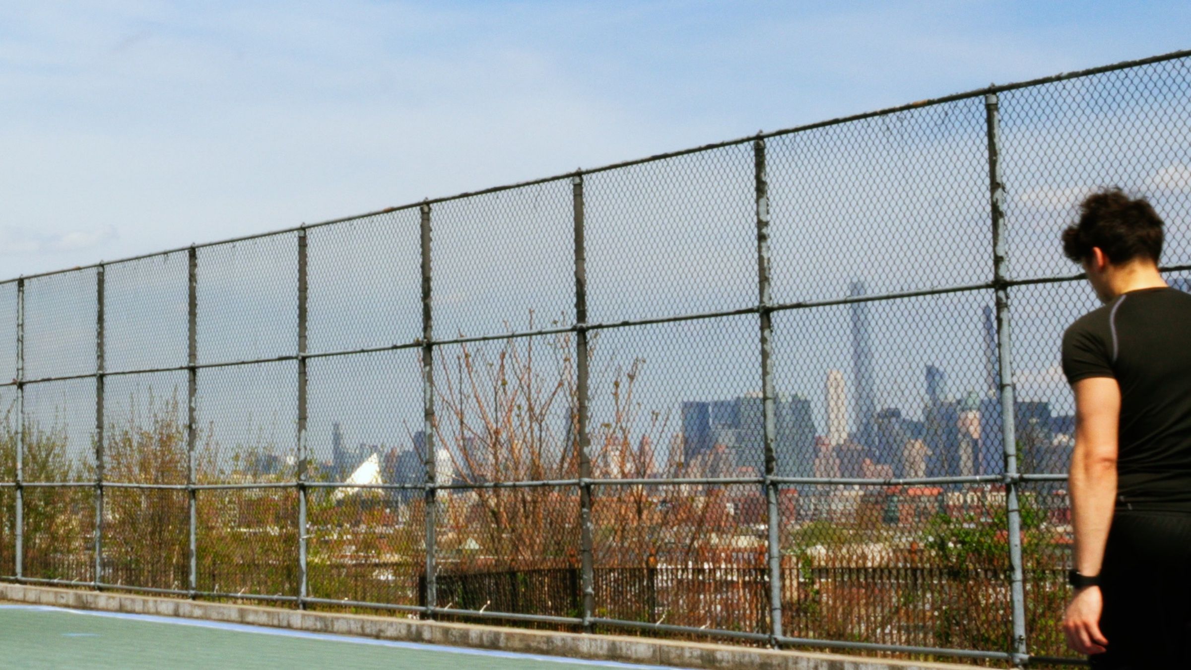 A player walks along the court fence, the Manhattan skyline beyond it.