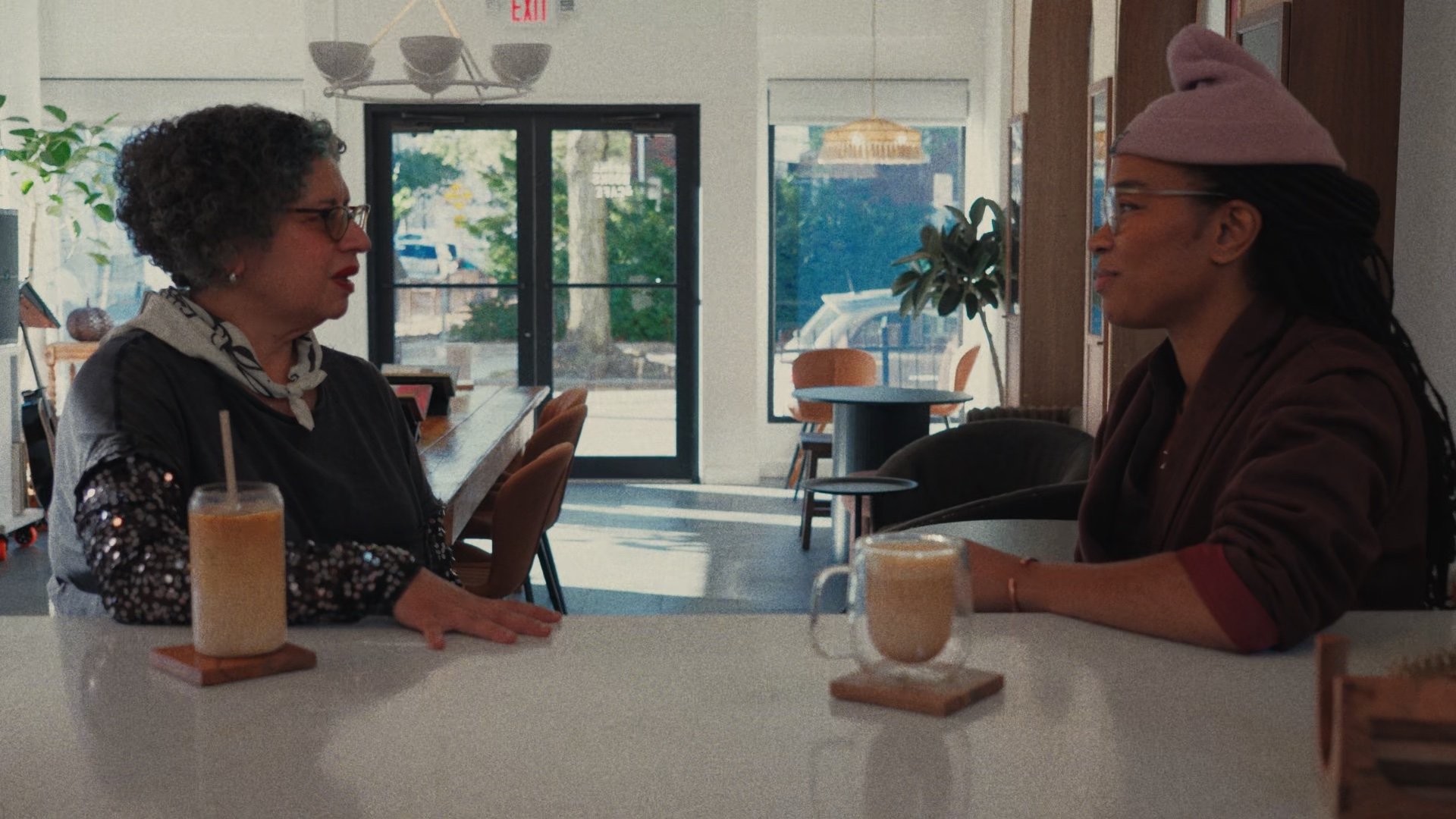 Two women in conversation at a white café table, morning light behind them.