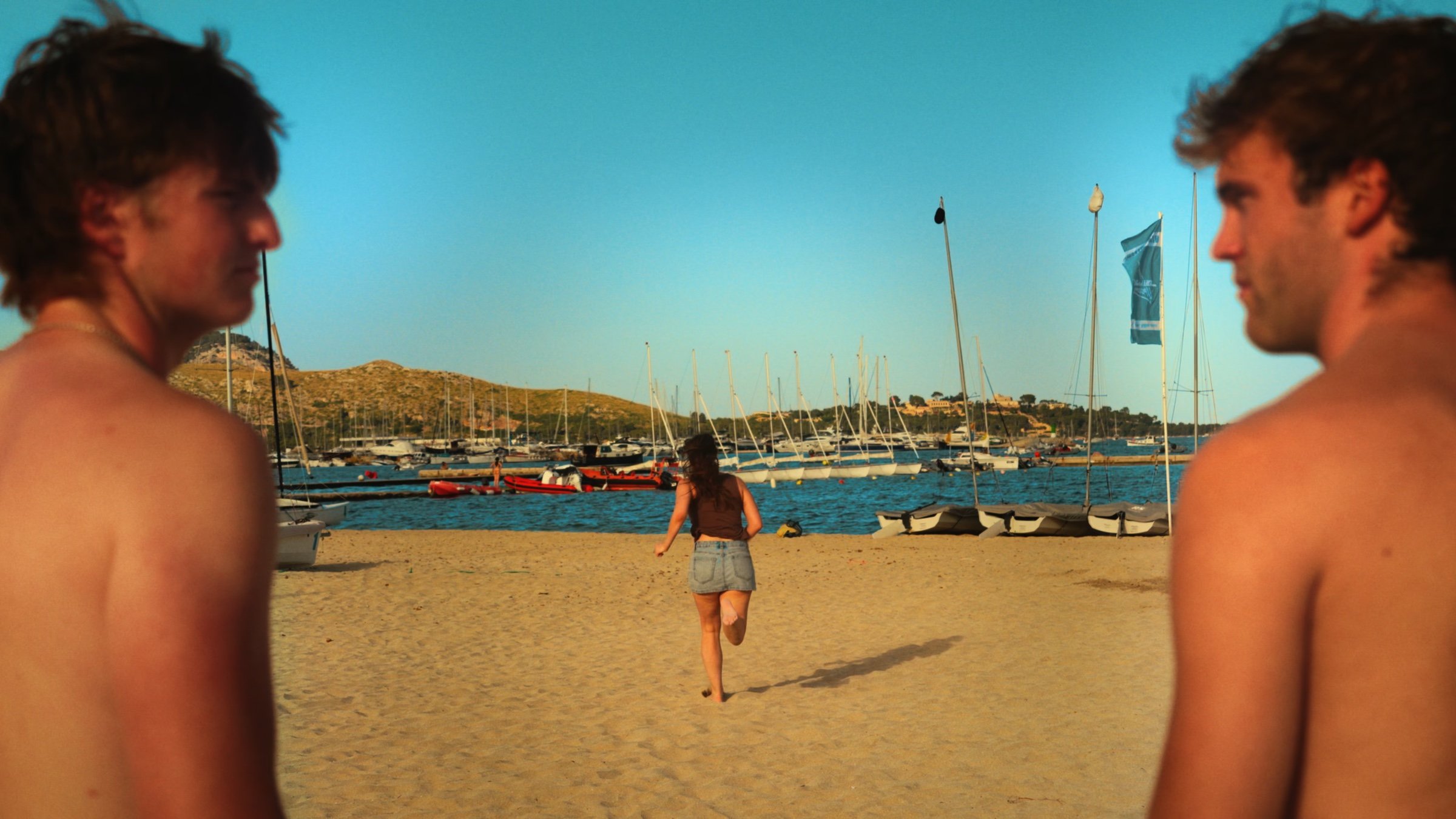 Two young men frame a woman running across the sand toward a harbour of sailboats.
