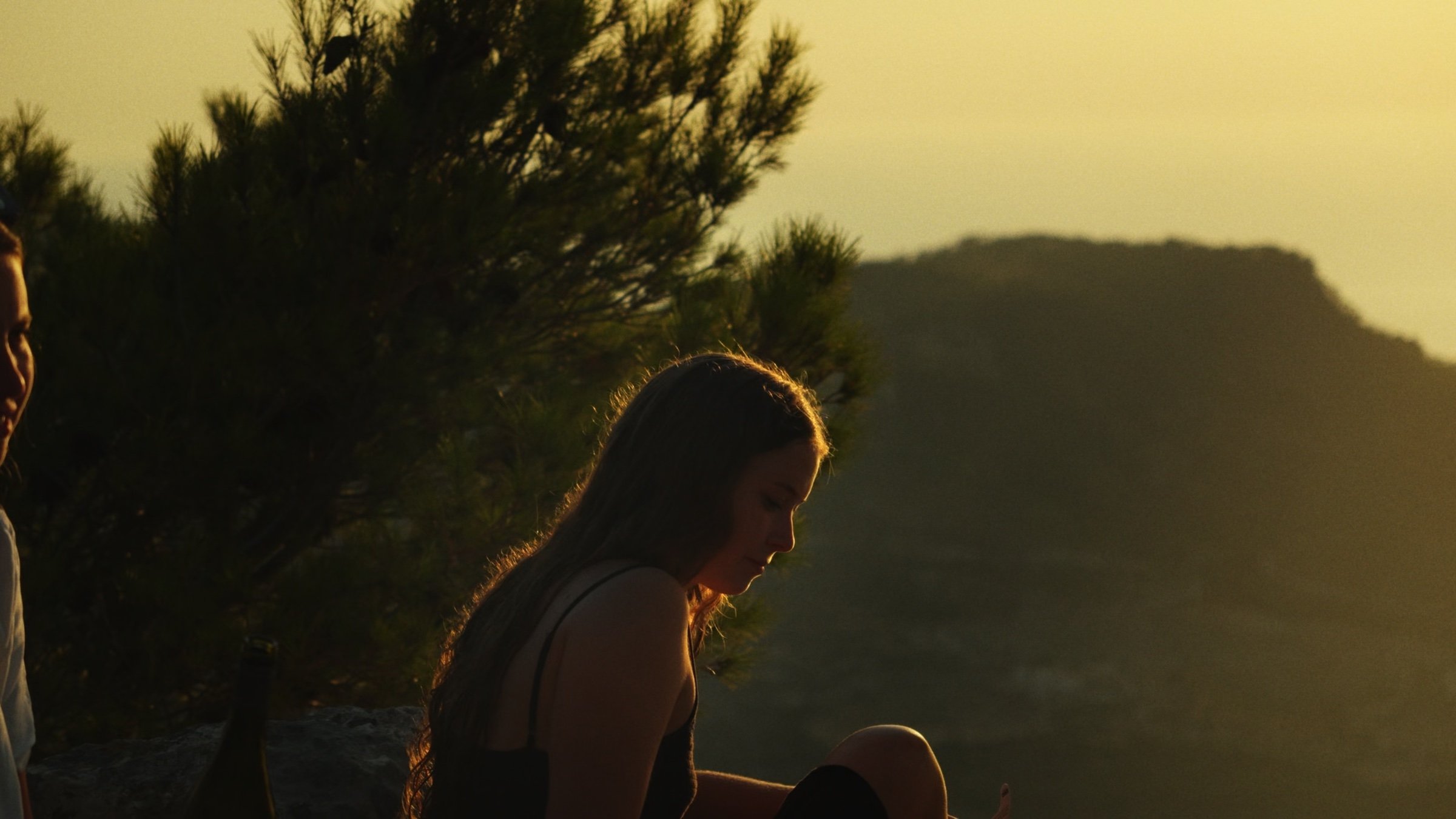 A young woman sits in golden evening light above the hills.