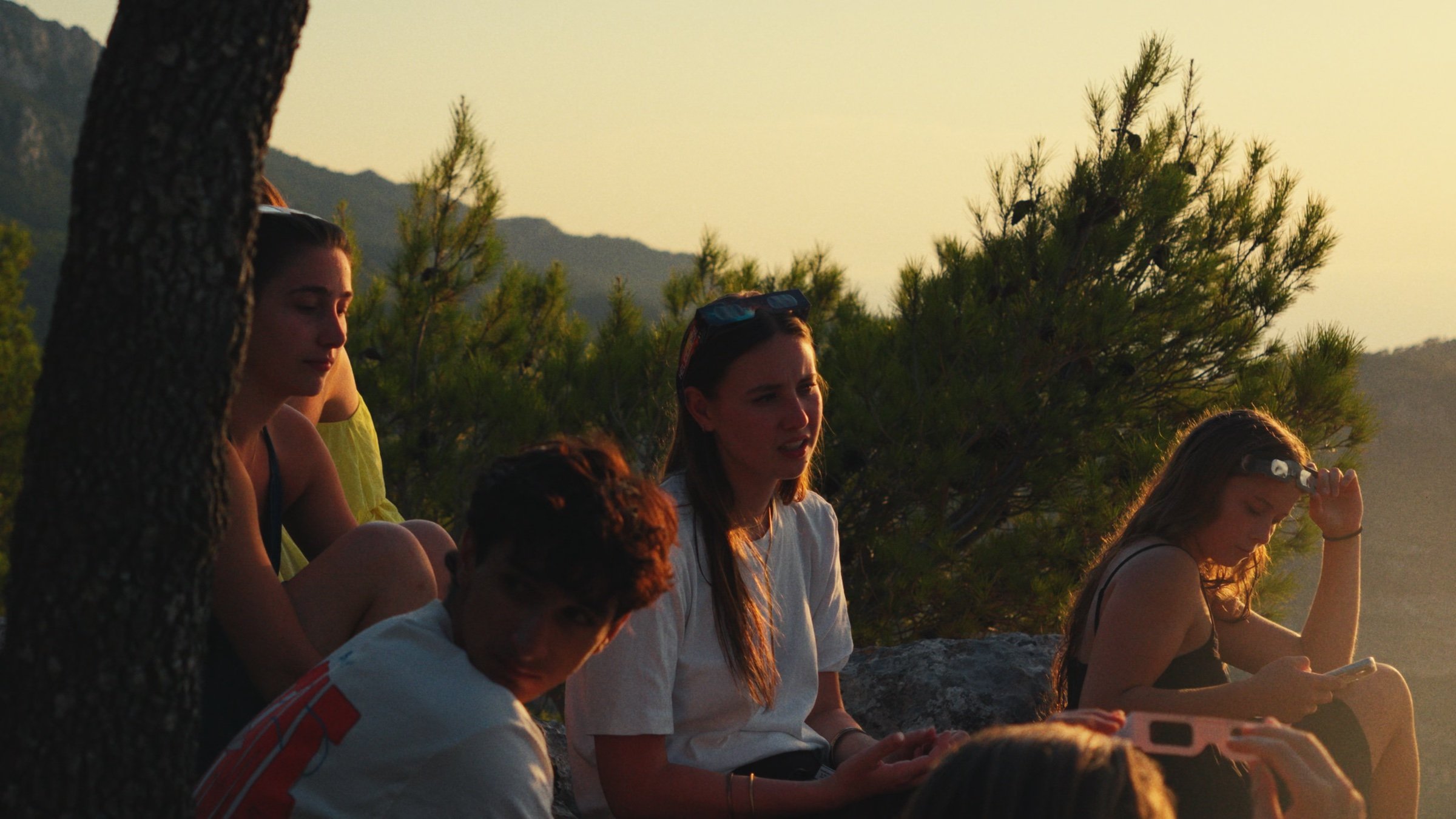 Friends gather on a pine-covered clifftop at sunset.
