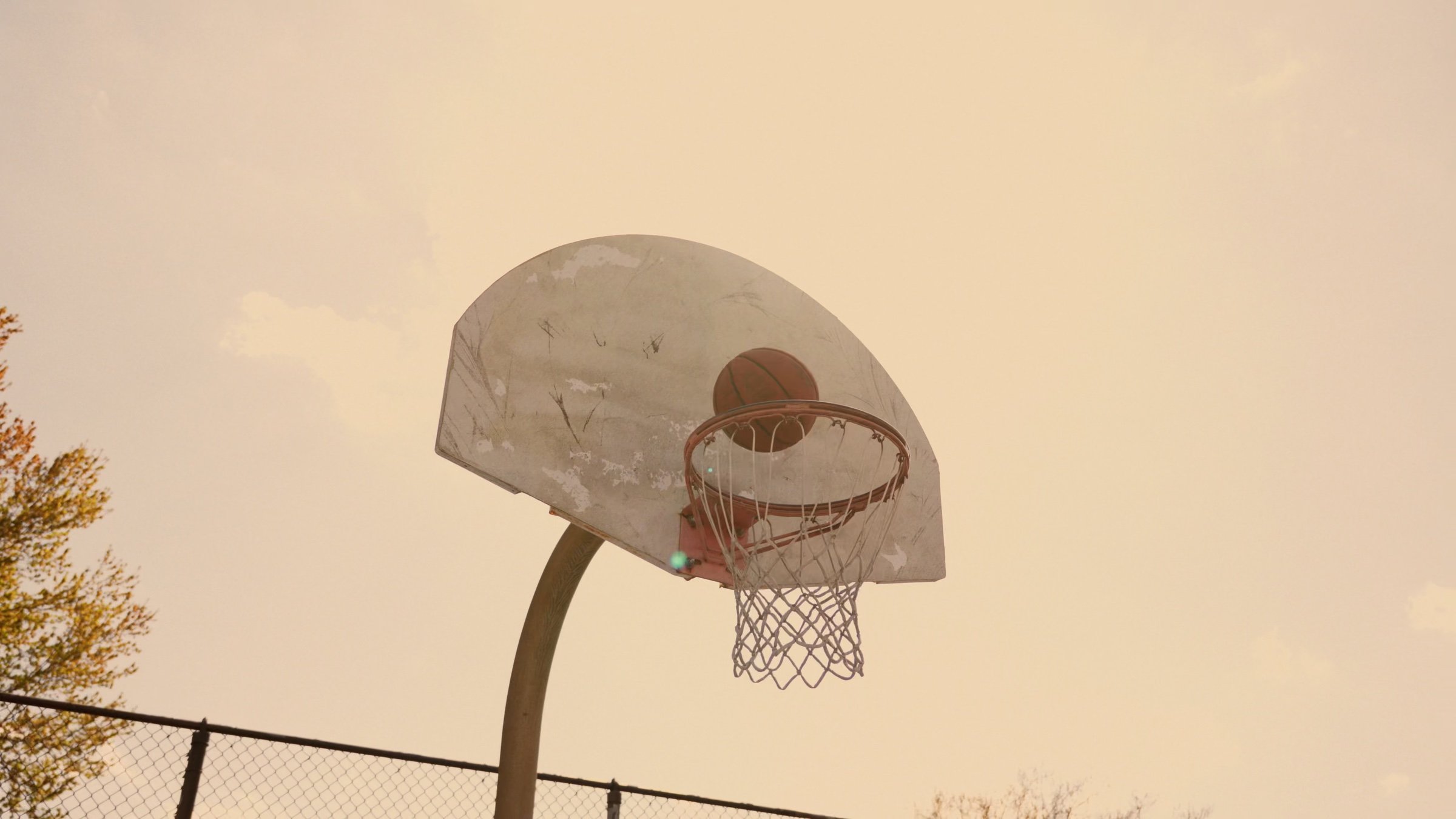 A basketball drops toward a weathered hoop against a warm evening sky.