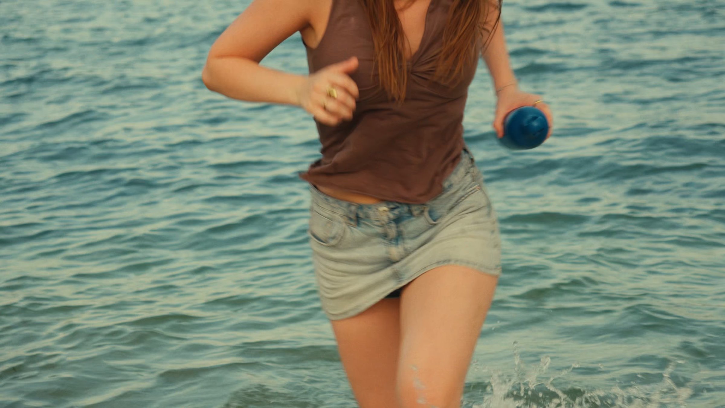 A woman wades into the sea holding a blue bottle.